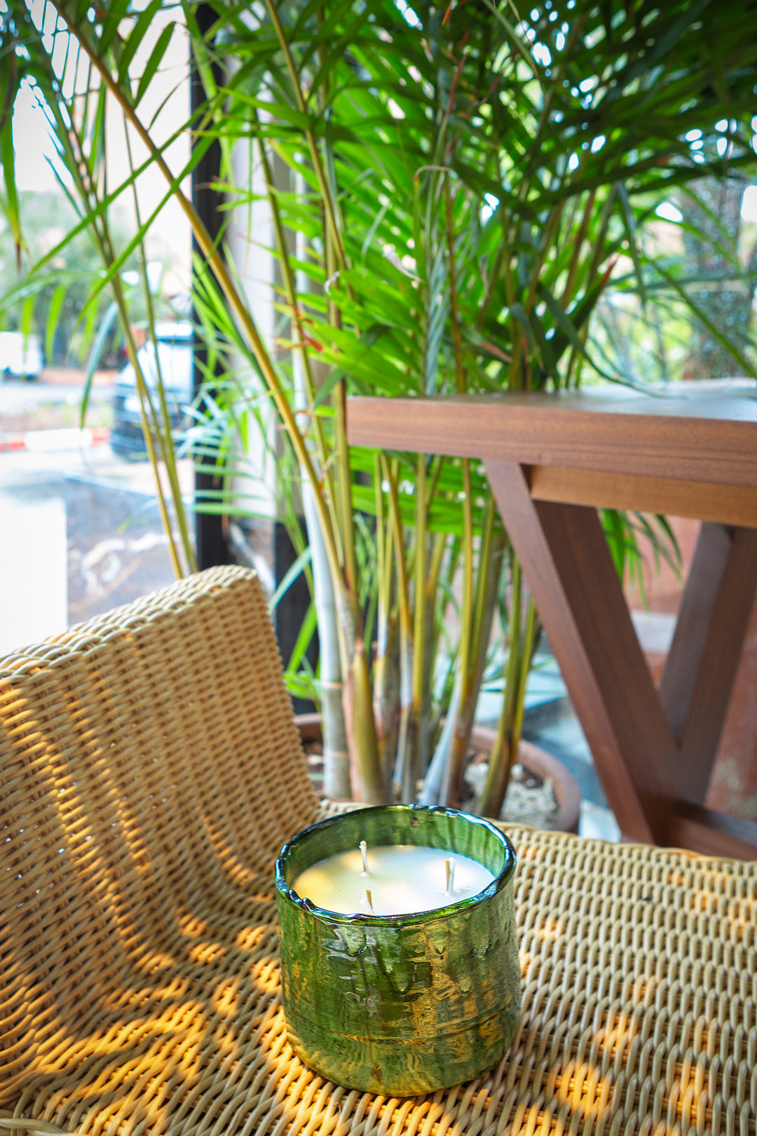 Green candle in a textured holder on a wicker chair with plants and a table in the background.