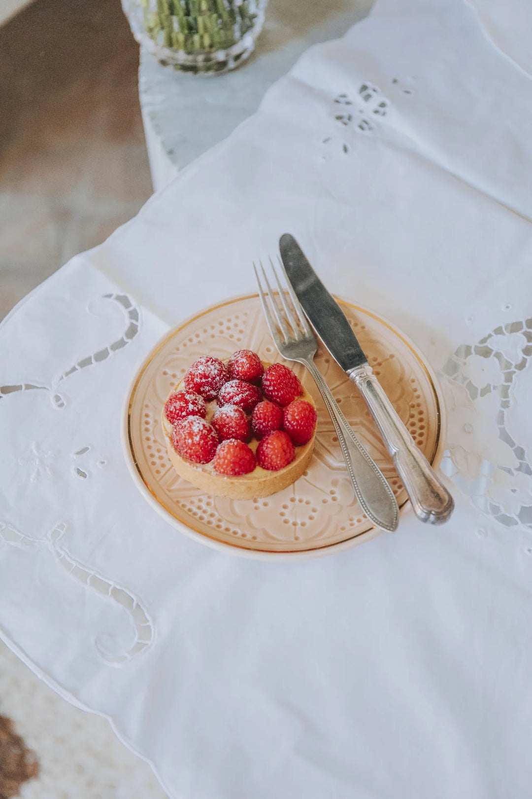 Small tart with raspberries on a decorative plate with a fork and knife on a white lace tablecloth.