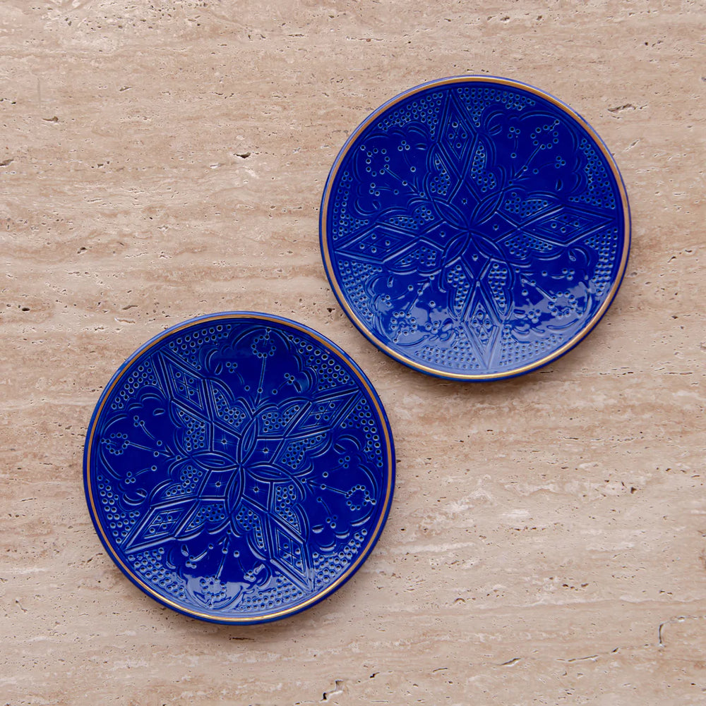 Two round, blue ceramic plates with gold engraved patterns, placed on a marble surface.